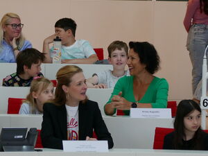 Kinder und Erwachsene sitzen im Plenarsaal. Einige Erwachsene haben Namensschilder auf den Tischen vor sich.