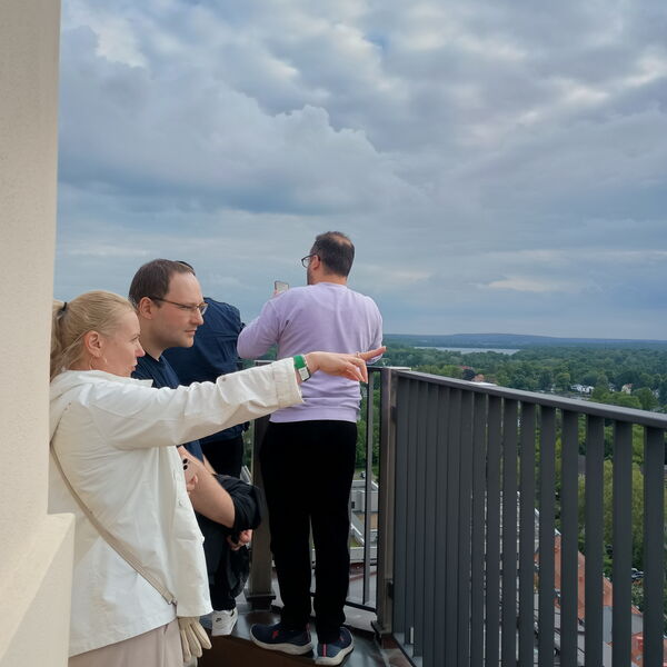 Menschen schauen von einer Aussichtsturm über Potsdam. Blonde Frau mit weißer Jacke streckt Arm aus und zeigt mit Finger nach rechts. Neben ihr ein Mann der in die gezeigte Richtung schaut.