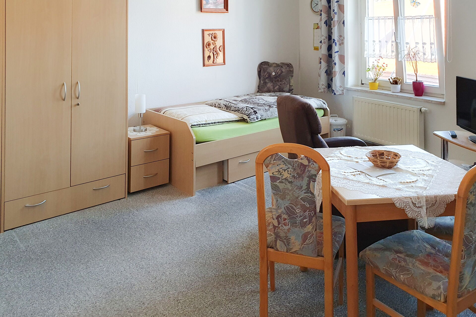Bewohnerzimmer- Links an der Wand ein Kleiderschrank, Nachttisch und Bett. Rechts: ein Fenster, ein Fernsehtischen mit TV und im Raum noch ein kleiner Tisch mit zwei Stühlen.
