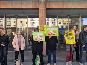 Auf dem Gehweg vor einem großen Gebäude mit vielen Fenstern stehen ein paar Menschen mit Schildern, die sie vor sich  halten.