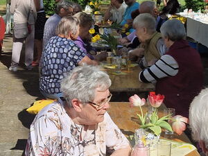 Biertischgarnituren, draußen, bei Sonne, Menschen sitzen an mit Getränken an Tischen. Rechts an der Wand eines Gebäudes sind Tische mit Deko zum Verkauf.