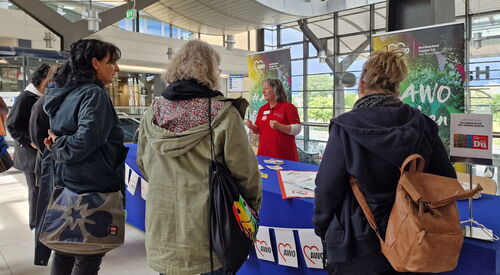 In den Bahnhofspassagen. Stand der AWO mit mehren Personen die sich informieren davor. Frau im roten AWO-T-Shirt steht hinter dem Tisch mit Info-Material.