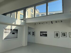 Großer Ausstellungsraum. Leer. Weiße Wände. Schwarz-weiß Bilder/Fotografien hängen an den Wänden. Oben befinden sich Fenster durch die, die Sonne scheint.