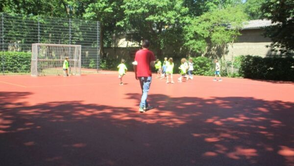 Sportplatz mit Tor. Torwart steht im Tor andere Kinder Fußballteams jagen dem Ball nach. Ein Erwachsener im roten T-Shirt betritt den Platz. Die Sonne scheint.