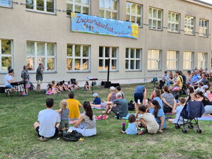 Ein Gebäude, davor eine Wiese. Viel Publikum sitzt auf der Wiese. Am Gebäude sitzen Kinder, ein Mann mit einem Keyboard und ein Mann hat ein Mikrofon in der Hand.