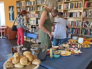 Ein Raum mit großem Bücherregal an der Wand. Im Raum ist ein Büfett aufgebaut. Brötchen, Käseteller, Wurstteller, halbe Eier, Honig und Marmelade, Obst, Kaffee Thermoskannen. 4 Menschen bedienen sich am Büfett.