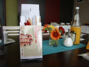 Auf einem Tisch steht ein Aufsteller mit Infomaterial zum Netzwerk, Postkarten und Stiften. Im Hintergrund sieht man Getränke und einen bunten Blumenstrauß.