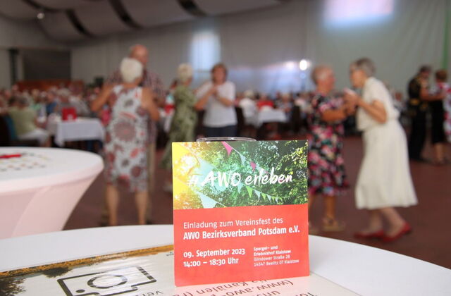 Einladung zum Vereinsfest steht an ein Glas gelehnt auf einem Tisch. Im Hintergrund fröhlich tanzende Menschen und lange Kaffeetafeln.
