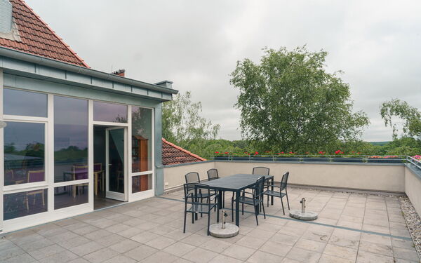 Große Dachterrasse mit Tisch, 6 Stühlen. Terrassentür steht offen, Glasfront.
