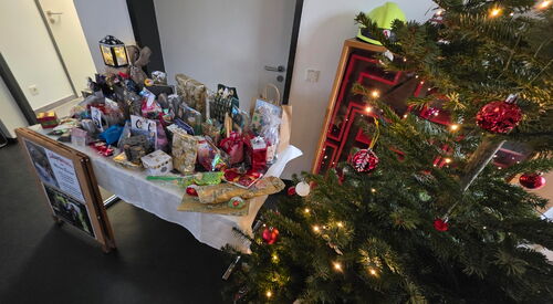 Links: Tisch mit Geschenken und Geschenttüten, rechts: geschmückter Weihnachtsbaum mit roten Kugeln und Lichterkette.