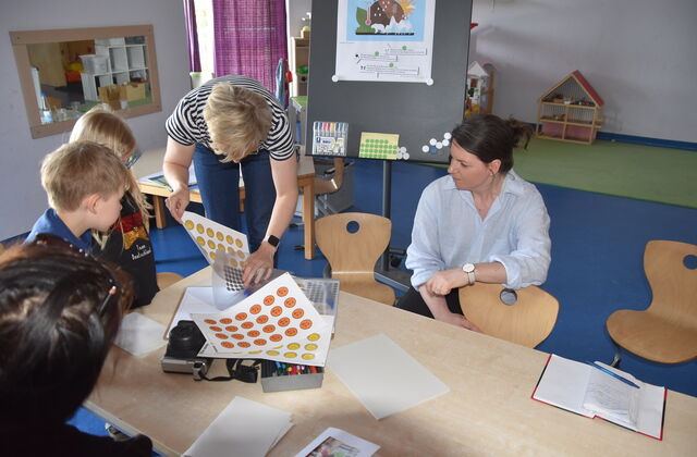 Zwei Kinder und drei Frauen am Tisch. Aufkleber von gelben und orangenen Smileys liegen auf dem Tisch. Frau mit kurzen blonden Haaren kramt noch mehr Smileys hervor. Frau mit schwarzen Haaren hockt am Tisch und stützt sich mit einem Arm auf Kinderstuhl.