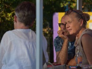 Ein Mann und eine Frau an einem Stehtisch. Mann mit Brille und T-Shirt. Frau mit blonden langen Haaren zum Dutt gebunden und Brille auf den Kopf geschoben. Beide schauen in die gleich Richtung.