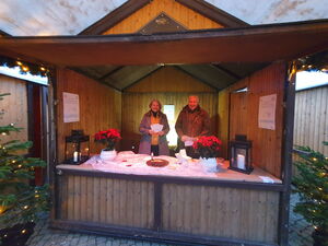 Weihnachtlicher Stand aus Holz mit Tannenbäumen rechts und links. Zwei Weihnachtssterne-Pflanzen und eine Frau und ein Mann im Stand.