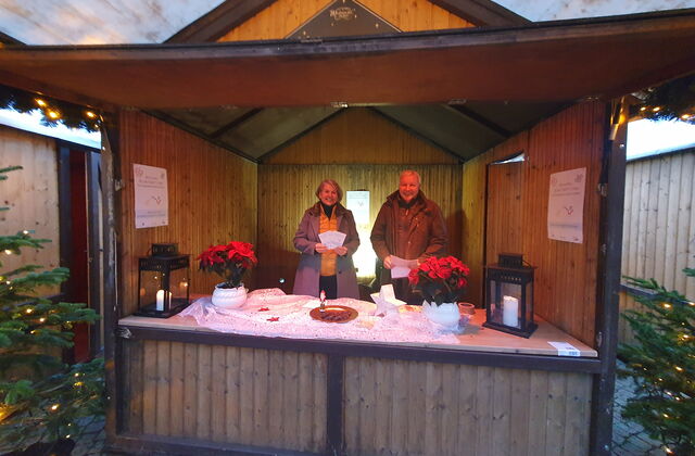 Weihnachtlicher Stand aus Holz mit Tannenbäumen rechts und links. Zwei Weihnachtssterne-Pflanzen und eine Frau und ein Mann im Stand.