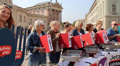 Auf dem Alten Markt in Potsdan. Die Sonne scheint. 6 Frauen und ein Mann stehen in einer Reihe und halten rote volle Aktenordner vor sich die das Logo: "Schule SATT" tragen.