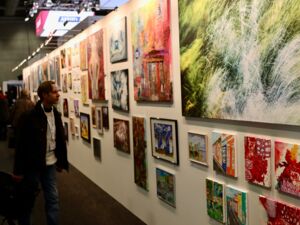 Ausstellungsraum. Viele Bilder an einer langen weißen Wand. Ein junger Mann betrachtet die Kunstwerke.