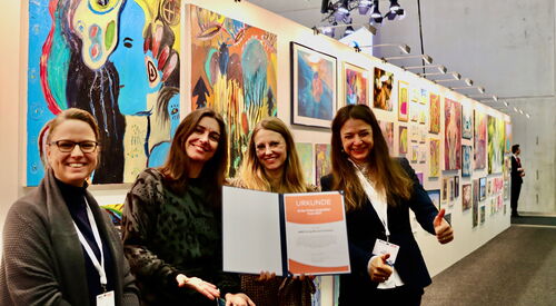 Vier junge Frauen sehr fröhlich und ausgelassen. Eine hält beide Daumen hoch. Sie halten die Urkunde / Auszeichnung in die Kamera. Sie stehen im Ausstellungsraum, hinter ihnen eine  lange Wand mit vielen Kunstwerken. Alle freue sich über die Urkunde.