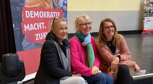 Drei Frauen sitzen auf einer Stufe / der Bühne vor einem AWO Roll-Up auf dem steht: Demokratie macht Zukunft. Die drei Frauen lächeln in eine Kamera.