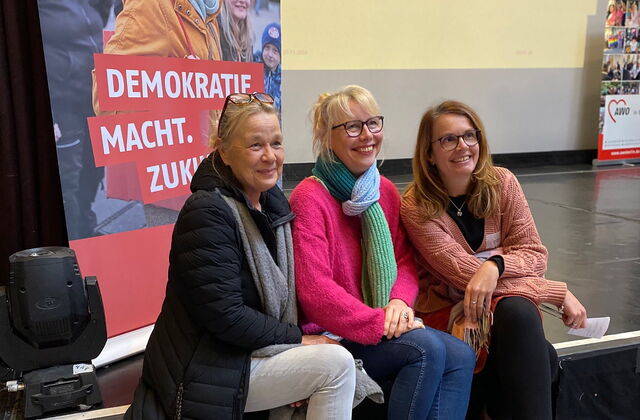 Drei Frauen sitzen auf einer Stufe / der Bühne vor einem AWO Roll-Up auf dem steht: Demokratie macht Zukunft. Die drei Frauen lächeln in eine Kamera.
