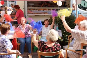 Seniorinnen sitzen in einem Stuhlkreis und schwenken farbige Tücher in der Luft. Die Tücher sind blau, lila, gelb und rot.