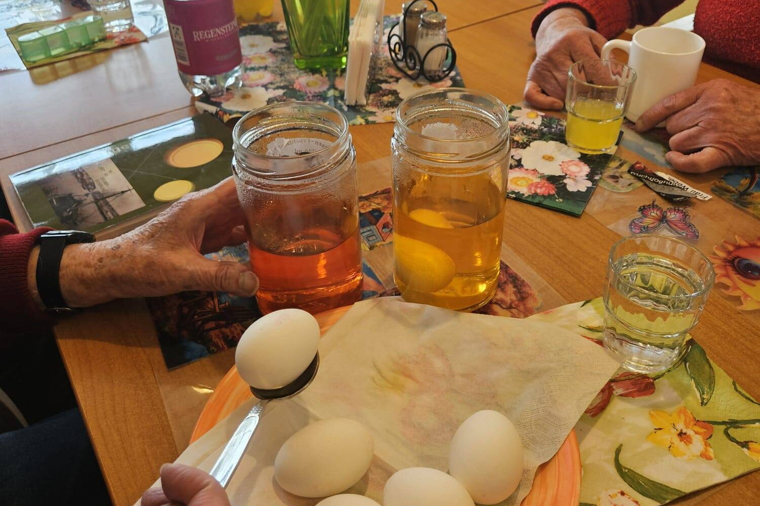 Eier färben. Einmachgläser mit farbiger Flüssigkeit. Hände von älterer Frau mit Esslöffel mit weißem Ei.