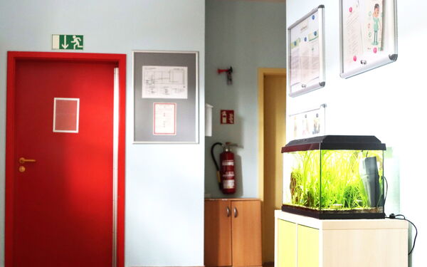 Ein Flur mit einer roten Tür. Über der Tür das grüne Notausgang-Zeichen. Ein Schränkchen mit einem Aquarium darauf. Über einem anderen Schränkchen in einer Ecke rechts von der roten Tür ist ein Feuerlöcher angebracht.