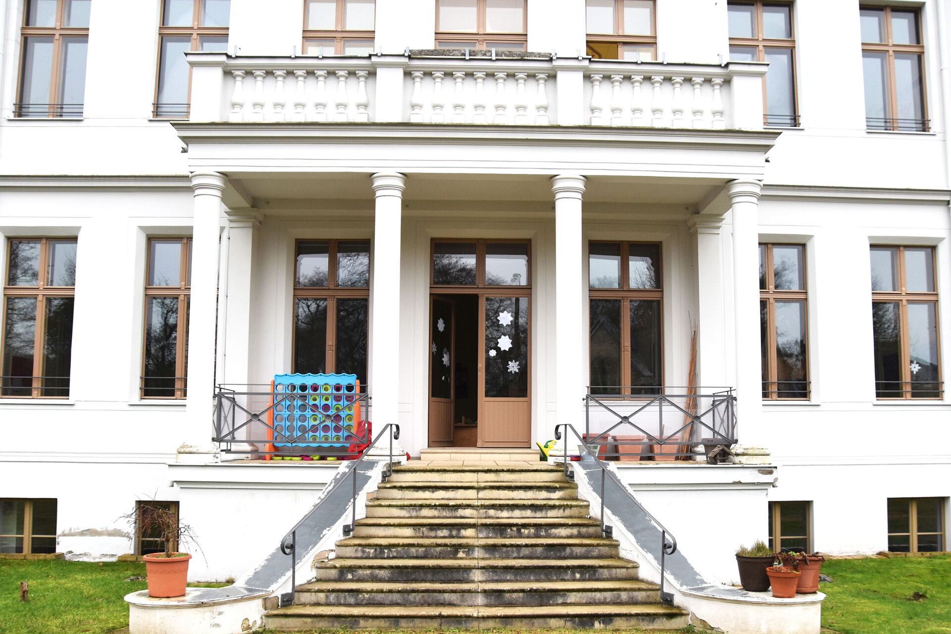 Eine Treppe führt hinauf zu der Eingangstür eines Gebäudes mit weißer Fassade und vier prächtigen Säulen. Die Eingangstür ist geöffnet.