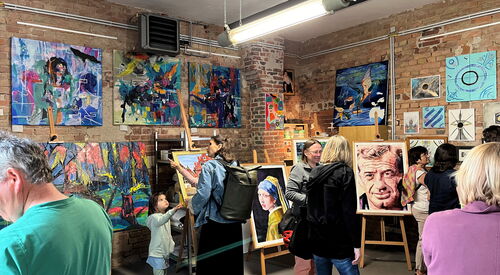 Menschen in einem Atelier, die sich Kunst Bilder, Gemälde ansehen. Es sind abstrakte Bilder aber auch Portraits darunter. Eine Frau im gelben Gewand und das Gesicht eines älteren Mannes mit Falten.