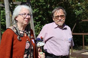 Portrait: Frau und Mann. Frau mit weißen vollen Haaren, sie trägt zwei geflochtene Zöpfe und eine große runde Brille, Mann, graue Haare, Bart, mit Brille mit getönten Gläsern. Er trägt ein kurzärmliges Hemd.