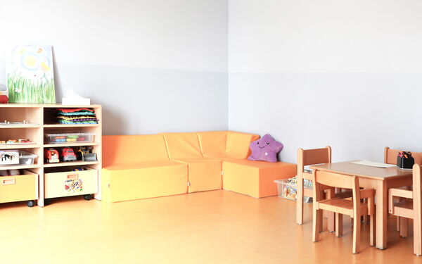 Freizeitraum. Freundlich, hell. An der Wand rechts ein kleiner Tisch mit 4 Stühlen. In der Eche eine gemütliche orangene Sitzecke mit einem lila Plüsch-Stern darauf. Daneben ein kleines Regal.