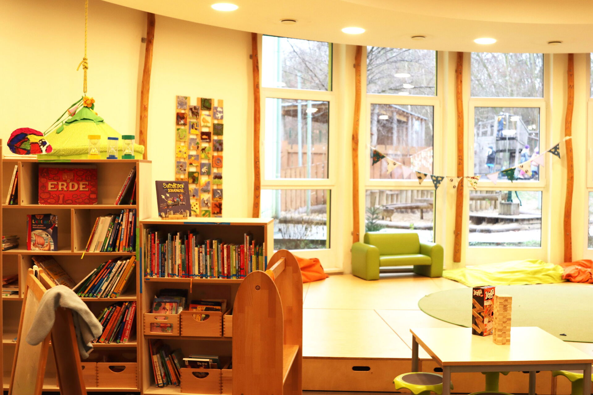 Freizeitraum, mit Regalen mit Büchern. bodengroßen Fenstern, Eine kleine grüne Couch steht vor einem Fenster. Vorne ist ein kleiner Tisch mit Hockern, auf dem Tisch ist ein Jenga Turm / Holz-Stapelspiel