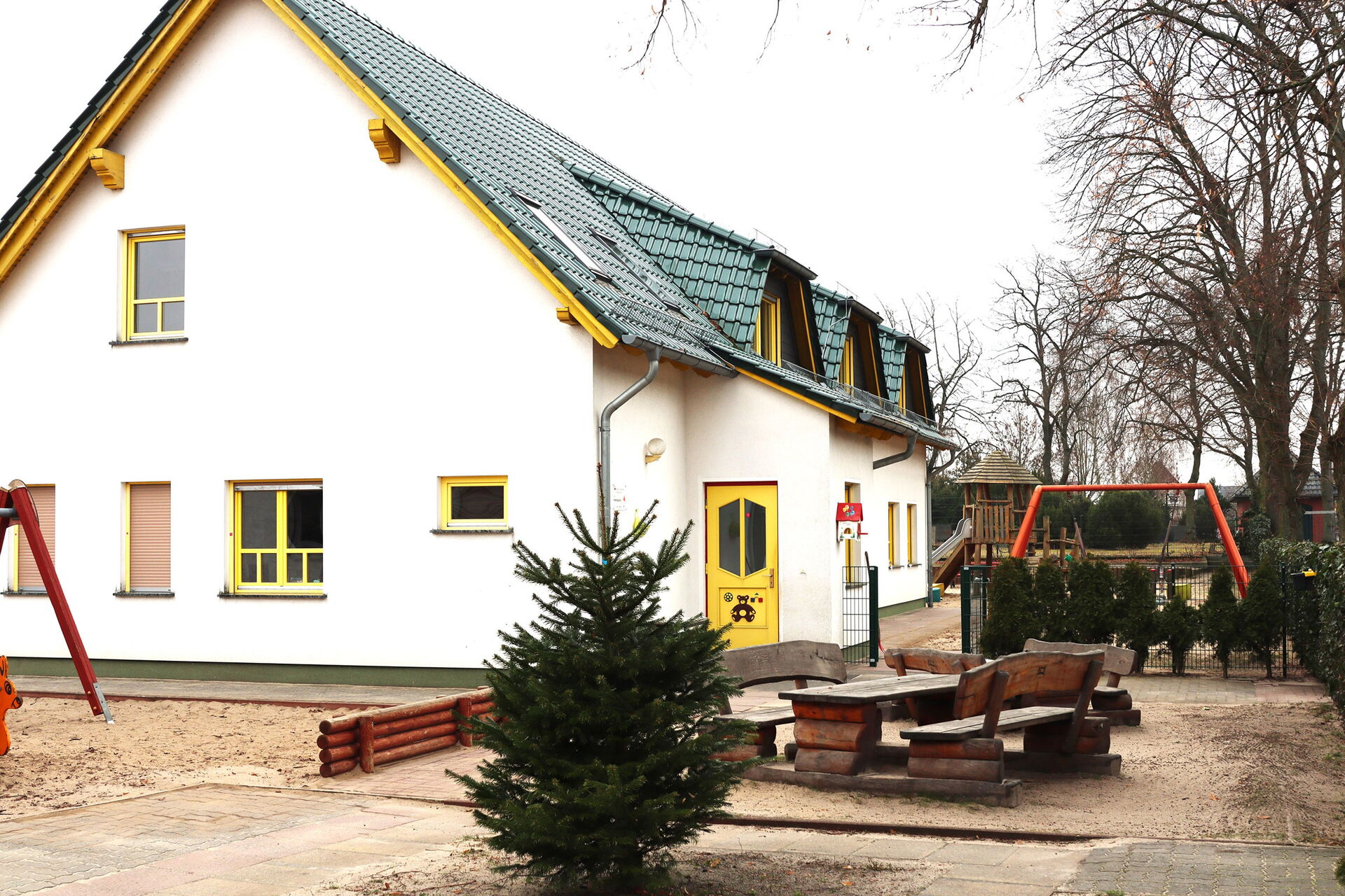 Weißes Haus mit gelber Tür und gelben Fensterrahmen, das Dach ist grün. Kahle Bäume, zwei Holzbänke mit Holztisch, ein kleiner Tannenbaum und Spielgeräte ums Haus verteil. Schaukel, Klettergerüst mit Rutsche.