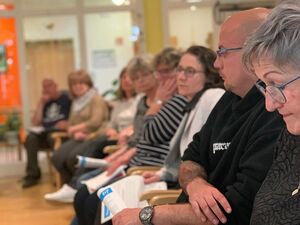 Großer Raum. 8 Menschen sitzen in einer Reihe. Stuhlreihe von der Seite fotografiert.