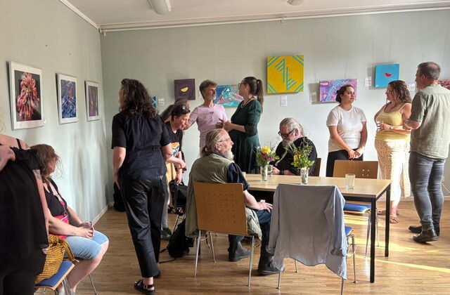 Ausstellung. Bilder hängen an weißen Wänden. Menschen sehen sich die Bilder an.