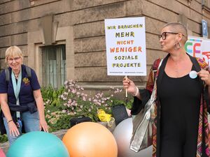 Farbige Pezzibälle und eine Frau die lächelnt bei den Bällen steht. Eine andere Frau mit sehr kurzen Haaren, im schwarzen Kleid hält ein Protestplakat vor sich darauf steht: Wir brauchen mehr nicht weniger soziale Projekte.