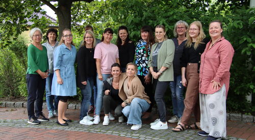 Draußen unter Bäumen. 11 Frauen stehen versetzt nebeneinander, in der Mitte sind zwei Frauen in die Hocke gegangen. Alle lächeln fröhlich in die Kamera.