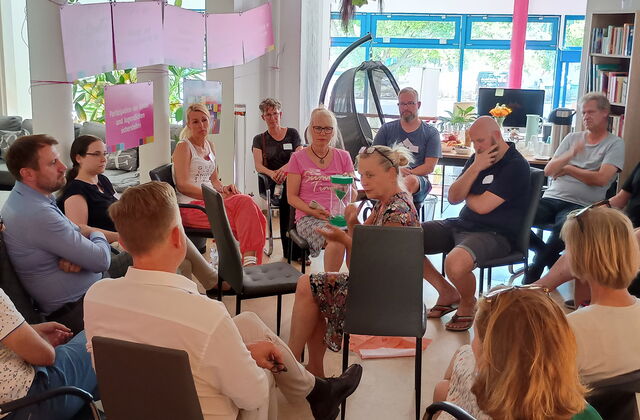 15 Menschen sitzen in zwei Stuhlkreisen. Frau mit mittleren Kreis hält eine grüne Sanduhr durch die grüner Sand rinnt.