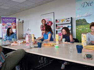 4 Frauen sitzen in einem Konferenzraum an einem großen Tisch. Eine Frau mit roten langen Haaren steht vor einer Tür und hält eine große Zuckertüte in den Armen.