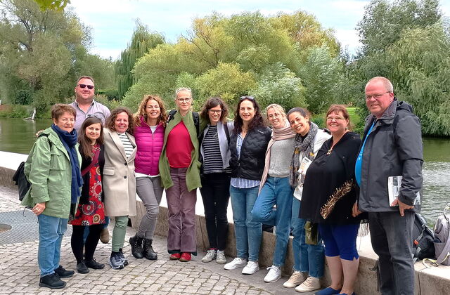 Gruppenbild. 10 Frauen, 2 Männer stehen an einem Fluss. Bäume und viel grün im Hintergrund auf der anderen Flussseite.
