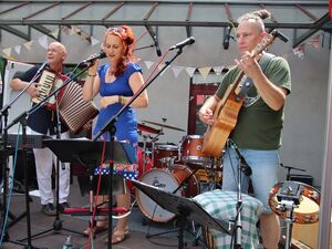 Eine Band auf der Bühne. Sängerin in der Mitte am Mikrofon, rechts der Gitarrist und links hinten ein älterer Mann mit Akkordeon. Die Frau trägt rote lange Haare und ein blaues Sommerkleid.