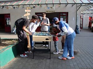 Draußen, Sonne scheint. Jugendliche und ein kleiner schwarzer Junge an einem Kicker. Sie spielen miteinander.