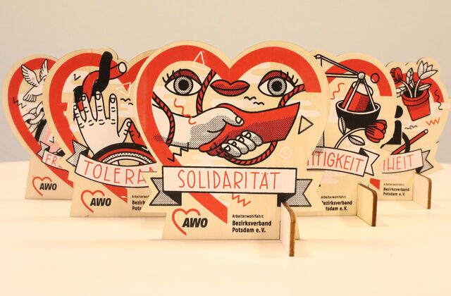 Tischaufsteller. In Herzform. Ganz vorn Solidarität, dahinter versetzt Toleranz und Gerechtigkeit. Dahinter sind noch zwei, die sind aber zum Teil verdeckt, durch die vorderen. Darauf abgebildet sind Symbole die zu den Aufschriften passen.