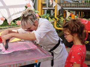 Eine Frau in weißer Kleidung, Sonnenbrille und blondem Haar beugt sich über einen Tisch mit rosa Farbe und hat ein Gerät in beiden Händen. Ein Mädchen guckt zu.