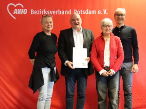 Portrait: 4 Personen vor roter AWO Wand. Frau, blonde lange Haare hochgesteckt, Brille auf den Kopf geschoben, Mann mit Glatze, weißem Hemd, schwarzem Sakko hält ein Papier vor sich, Frau mit weißen Haaren und Brille und großer schlanker Mann mit Brille.