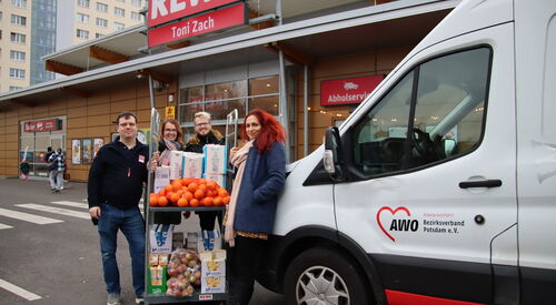 Vor einem REWE Markt. Ein weißer Transporter der AWO steht davor. Eine Frau mit langen wilden roten Haaren lehnt am Transporter. 3 weitere Personen schieben einen Rollbehälter beladen mit Lebensmitteln. Alle lächeln in die Kamera.