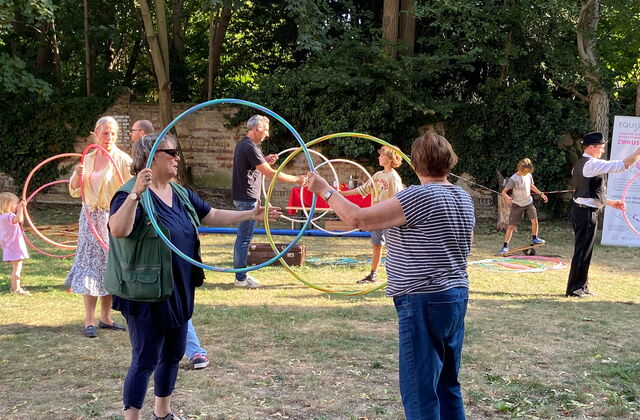 Jeweils zwei Personen halten zwei Hula Hoop Reifen senkrecht in den Händen. Mehrere Paare aus groß und klein stehen sich so gegenüber.