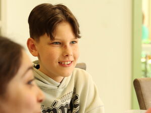 Junge am Tisch, lächelt. Er hat einen vollen Teller mit Nudeln und Tomatensouce vor sich.