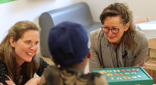2 Frauen sitzen an einen Tisch mit einer grünen Box. Auf der Bux steht "Wunsch-Essen-Kinder"  Mit dem Rücken zu Kamera ein Kind mit blauem Cappy.