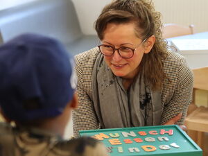 2 Frauen sitzen an einen Tisch mit einer grünen Box. Auf der Bux steht "Wunsch-Essen-Kinder"  Mit dem Rücken zu Kamera ein Kind mit blauem Cappy.