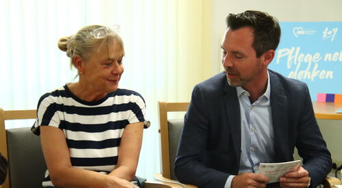 Frau mit blonden hochgesteckten Haaren und gestreiftem T-Shirt sitzt neben Mann in Anzug und Hemd, sie unterhalten sich.